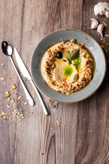 Hummus in a grey bowl seasoned with sunflower and pumpkin seeds, sesame, olives and olive oil. Close-up on a table with cutlery and seeds by side. Top view isolated on wooden background. Copy space.