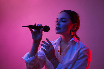 Female singer in neon light. Girl in white shirt with microphone. Concept of art, human emotions, music.