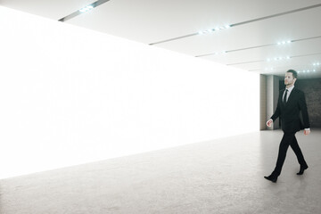 Young businessman in black suit walking in empty spacious hall room with blank white glowing wall...
