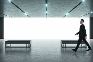 Obraz premium Businessman in black suit walking in a modern exhibition room with blank projection screen and benches. Education and presentation concept. Mock up
