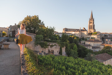A young blonde tourist enjoying her holiday and vineyard view of the Monolithic Church and village of Saint-Emilion in Bordeaux wine country on a sunny summer day.