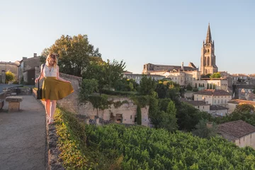 Fotobehang A young blonde tourist enjoying her holiday and vineyard view of the Monolithic Church and village of Saint-Emilion in Bordeaux wine country on a sunny summer day. © Stephen