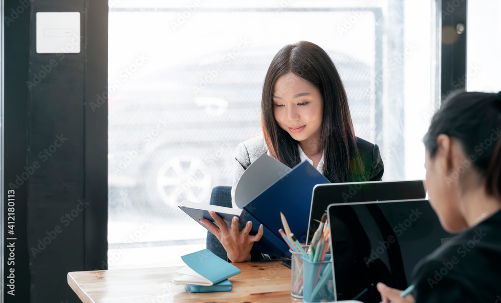 Wall mural young beautiful asian woman reading a book while sitting at office desk with her friend.