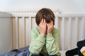 Close up portrait of a young boy of ten years old sitting alone at home and crying. Upset depressed child closing face by hands.