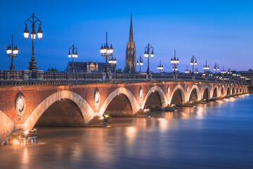 The historic Pont de Perre illuminated at night over the River Gardonne in Bordeaux, France with Bordeaux Cathedral in the background.