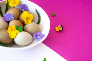 Easter eggs with yellow and lilac flowers in a white bowl on a white and pink background.