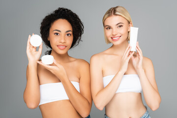 Smiling multiethnic women holding tube and jar with cosmetic cream isolated on grey