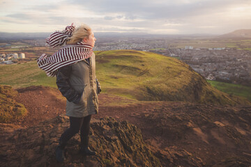 A young blonde female tourist with a scarf looks out to a cityscape view of Edinburgh, Scotland on a windy afternoon up Arthur's Seat.