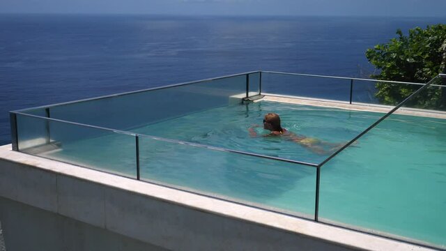 Happy middle aged woman swimming in an infinity pool overlooking an ocean. Luxurious resorts concept.