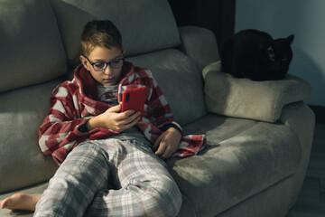 Boy wearing glasses and pajamas sitting on the sofa using his smartphone with a red phone case