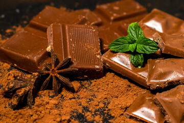 Different pieces of dark chocolate and cocoa powder on a dark surface. Mint leaves and star anise seeds complete the picture. Space for text
