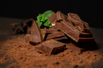 Different pieces of dark chocolate and cocoa powder on a dark surface. Mint leaves and star anise seeds complete the picture. Space for text