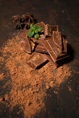 Different pieces of dark chocolate and cocoa powder on a dark surface. Mint leaves and star anise seeds complete the picture. Space for text