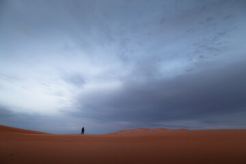 A distant figure exploring sand dunes desert landscape against a moody, dramatic sky at Erg Chebbi near the village of Merzouga, Morocco.