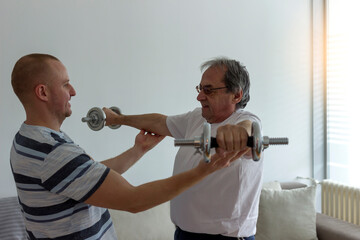 Shot of a friendly physiotherapist helping senior patient work out with weights at home. Senior man working out. Physiotherapist helping his mature patient to use dumbbells in a rehabilitation center.