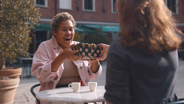 Two diverse female best friends spending time together sitting with gift on terrace in cafe