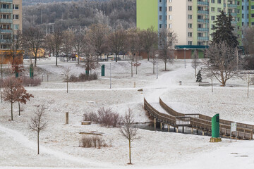 Snowy park in the city of Brno in the Czech Republic in the Novy Liskovec district. You can see prefabricated houses, a wooden bridge over the pond and climbing frames for children.