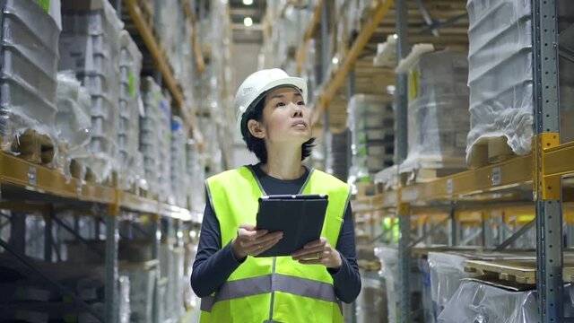 Young asian woman using tablet while walking in warehouse of modern factory. Front view of female employee doing work in storehouse, holding digital device in hand and looking at screen. Worker