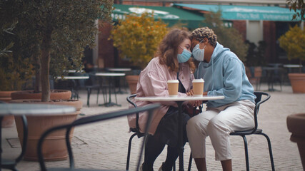 Fototapeta na wymiar Diverse lesbian couple in safety mask sitting at table in street cafe and hugging