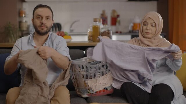 Muslim Husband And Wife, Who Cannot Take Their Eyes Off The TV, Fold The Dried Laundry. Home Laundry Folding Concept.