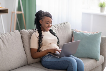 Obraz premium Joyful african lady sitting on couch at home, using laptop