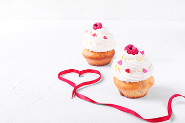 Cupcakes topped with whipped cream, heart shaped sprinkles and raspberry. Red satin ribbon forming heart on the white table