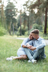 Naklejka na ściany i meble Outdoor shot of young happy couple in love sitting on grass on nature. Man and woman hugging, sunlight in summer park. Happy family in the evening sun light. The concept holiday