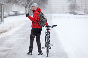 Young bicyclist showing thumb up while looking backward, man with bike standing on snowy lane at winter season