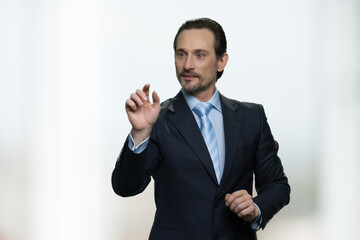 Mature businessman is touching invisible virtual screen. Wearing formal suit. White blurred background.