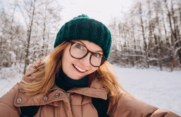 Young beautiful happy girl in green winter knitted hat takes selfie in winter forest. Looking at camera and smile. Travel and active life concept. Outdoors