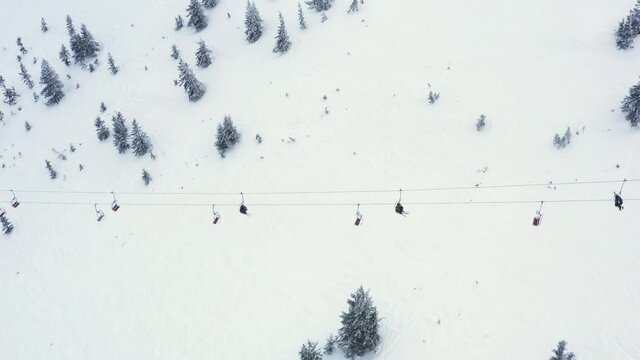 Ski lift elevator transporting skiers and snowboarders on snowy winter slope at mountain resort. Top view from drone chairlift moving on cable way in ski resort. UHD 4k video