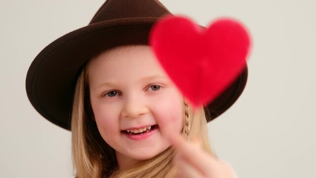 Little girl child in hat holding sweet heart shaped candy and eating licks. Valentine's day and share love concept, selective focus.
