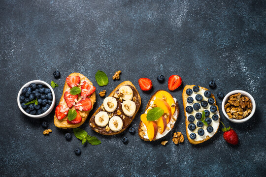 Sweet toast assortment. Chocolate banana, cream cheese and peach, peanut butter and strawberry, cream cheese and blueberry toasts. Black stone background, top view.