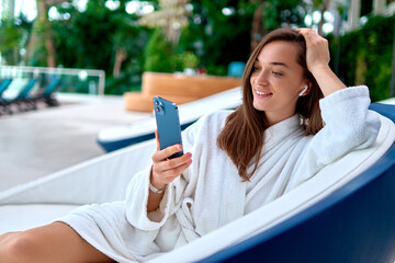Young attractive smiling woman wearing white bathrobe using smartphone for watching video and...