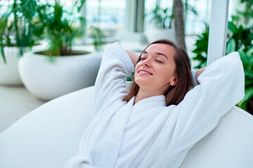 Serene calm woman wearing white bathrobe with closed eyes and hands behind head enjoying relaxing time and feeling good at a wellness spa resort. Easy lifestyle and satisfaction - obrazy, fototapety, plakaty