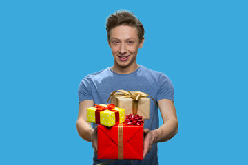 Portrait of smiling boy in t-shirt holding gift boxes. Positive schoolboy isolated on blue background.