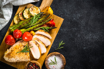 Grill menu, bbq dish. Chicken breast grilled with vegetables on a wooden serving board. Top view with copy space on black stone table.