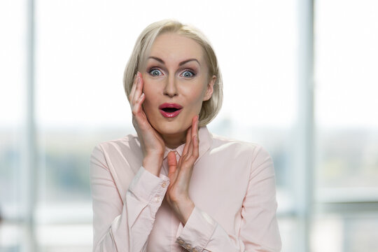 Happy and surprised mature businesswoman. Astonished american caucasian blond woman is touching her face. Blurred windows interior on the background.