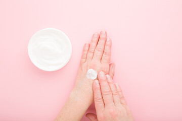 Mature woman hands using white anti aging moisturizing cream on light pink table background. Pastel color. Care about soft body skin in old ages. Daily beauty product. Closeup. Top down view.