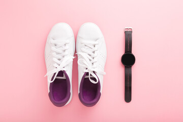 White female sport shoes and black smartwatch for walking, running or fitness on light pink table background. Pastel color. Closeup. Top down view.
