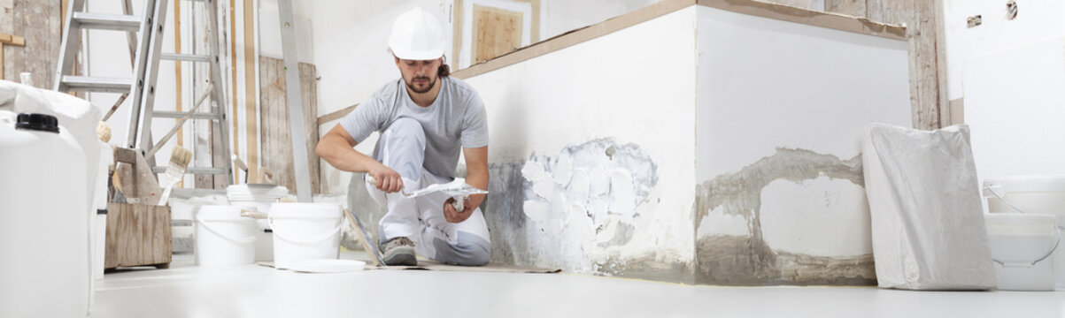 plasterer man at work, take the mortar from the bucket with trowel to plastering the wall of interior construction house site and wear helmet, panoramic image