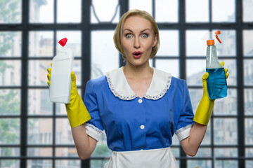 Surprised cleaning lady is raising her arms. Middle-aged blonde woman in housemaid's uniform is holding cleansers. Big window on the background.