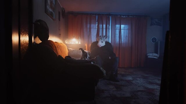 Eerie scene of a scary, masked stranger sitting in a dark living room, petting a cat. Mysterious man in an animal mask, goat costume. Devil waiting in the night, dark, unsettling scene.