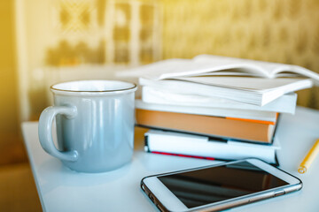 A stack of books, a mug of tea, and a mobile phone on a white table. A bookcase with books in the background in a modern interior. The concept of training and education.
