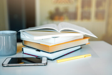 A stack of books, a mug of tea, and a mobile phone on a white table. A bookcase with books in the background in a modern interior. The concept of training and education.