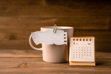 A piece of paper with a place for text is held by a paper clip to a mug, next to a calendar on a wooden table. Creative background for notes
