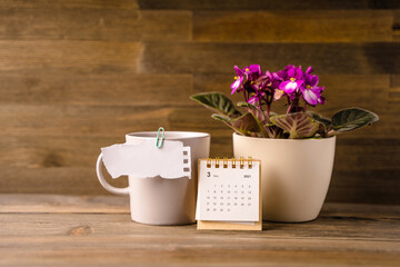 A piece of paper with a place for text is held by a paper clip to a mug, next to a calendar on a wooden table. Creative background for notes