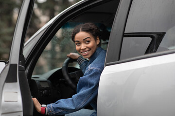Friendly smiling millennial african american female driver opens door and looks for fellow traveler