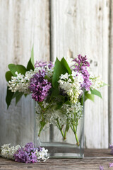 Fototapeta na wymiar A bouquet of spring purple and white lilacs in a vase on a light wood background. Glass transparent container with water. Rustic aromatic composition. Soft focus