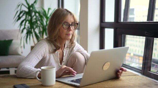 Woman using laptop at table and spending time at home during pandemic. Middle aged female looking at computer screen and searching on internet while sitting at desk in light room. American beautiful